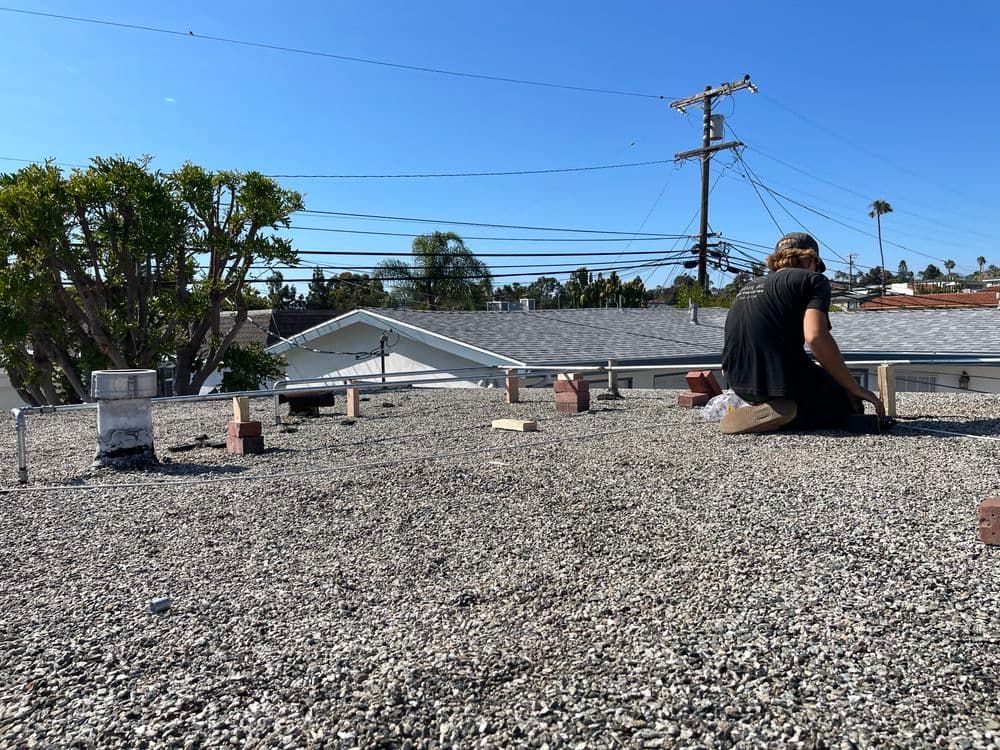 Roofer working on a gravel flat roof under a clear blue sky with power lines in view.