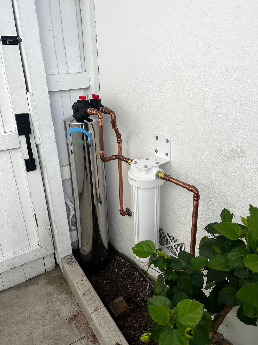 Shiny silver water filtration system with copper pipes next to a plant and wall.