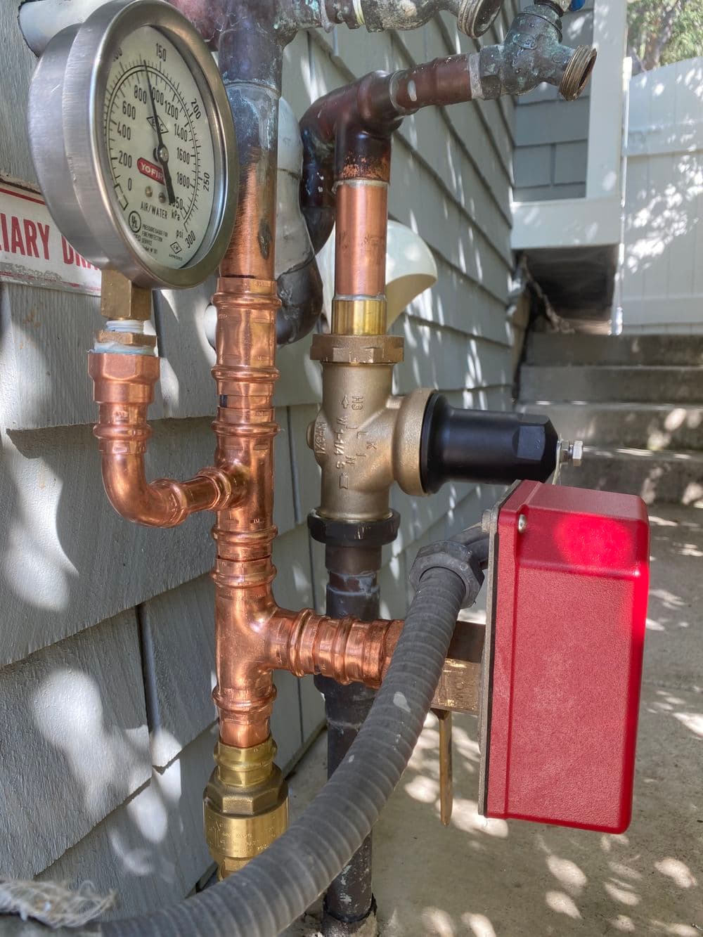 Close-up of a water meter and valve with copper pipes and red control box on a house exterior.