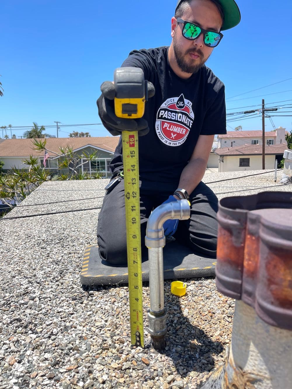 Plumber measuring pipe height on residential roof under clear blue sky.