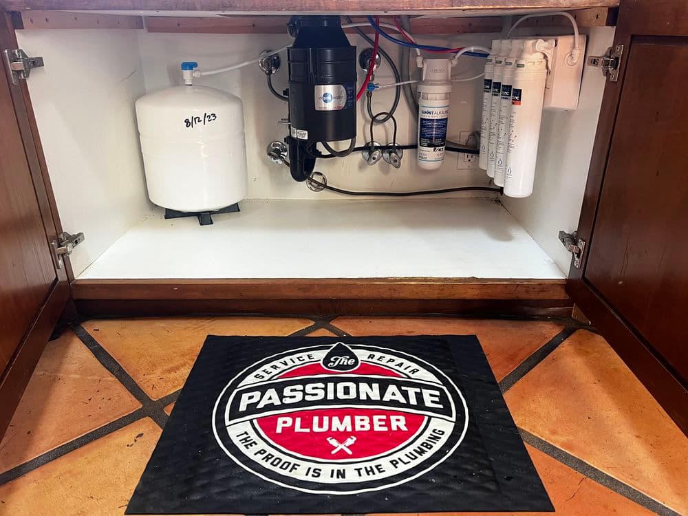 Under-sink plumbing setup with water filtration system and branded mat of The Passionate Plumber.