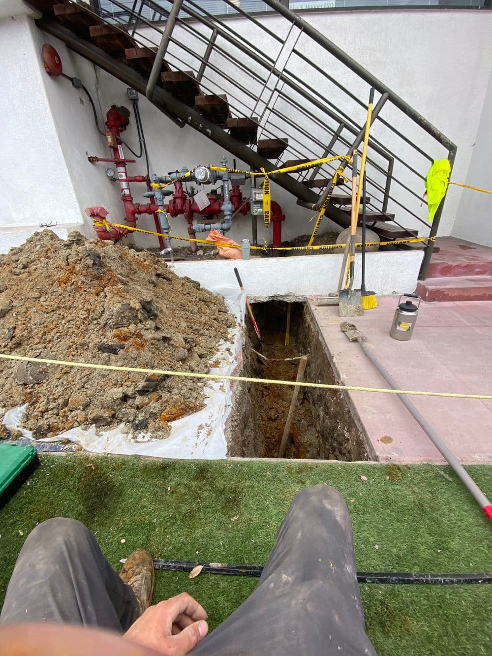 Construction site featuring an open trench near a staircase and utilities, with safety markers.