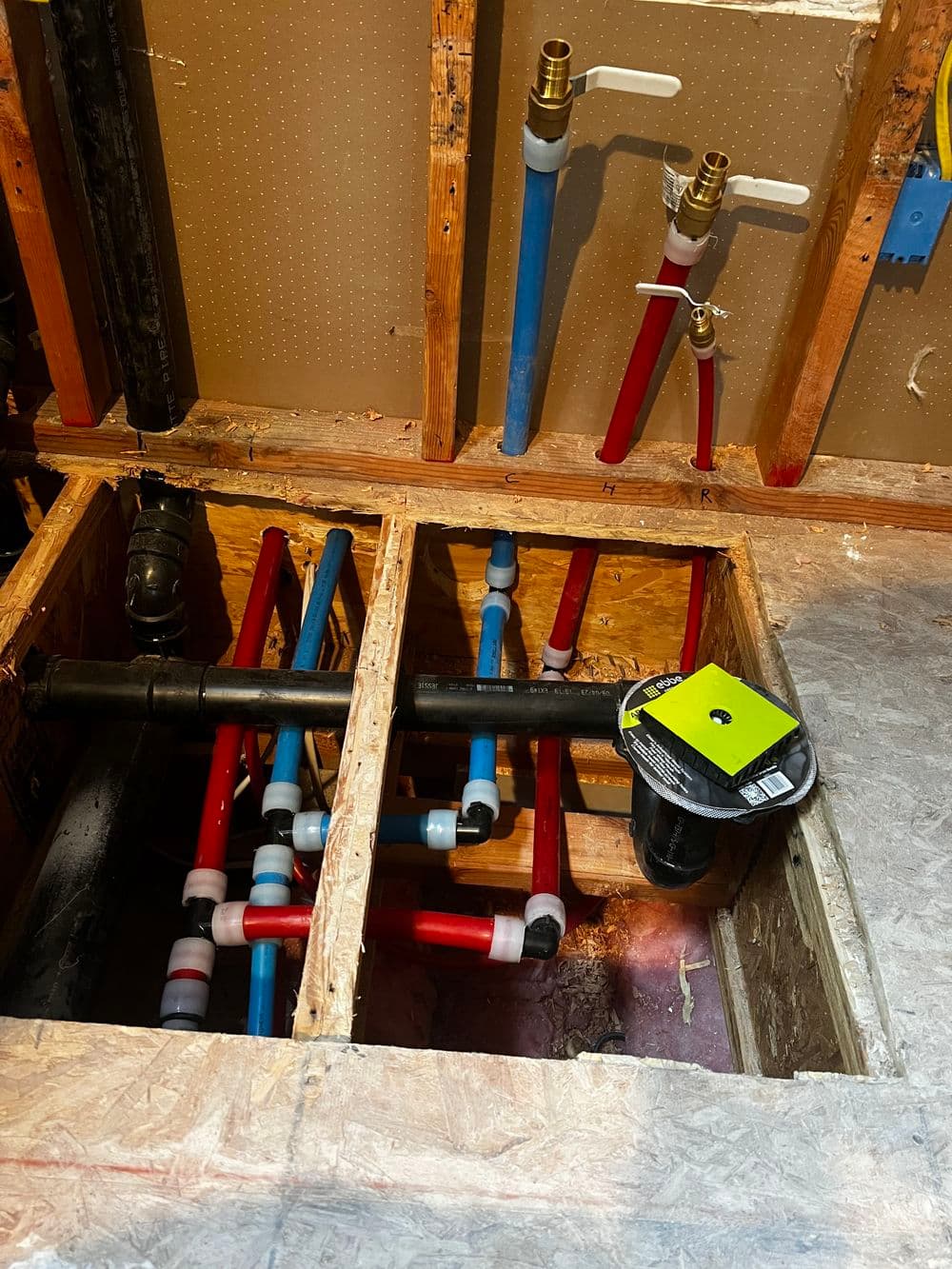 Plumbing installation with red and blue pipes in a crawl space under floorboards.