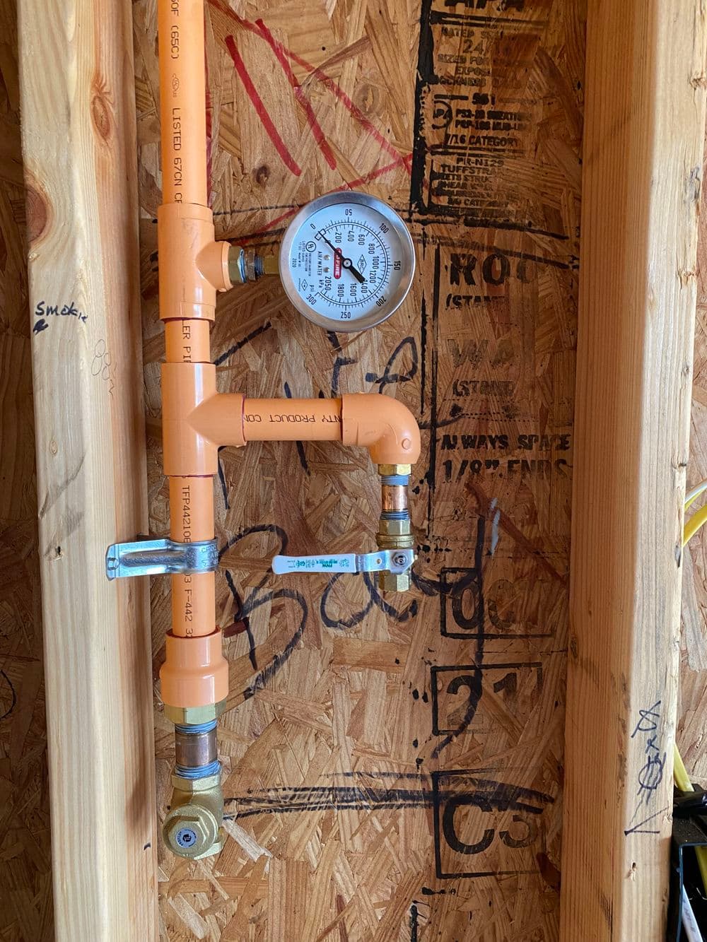 Orange PVC pressure gauge mounted on wooden wall with plumbing components and markings.