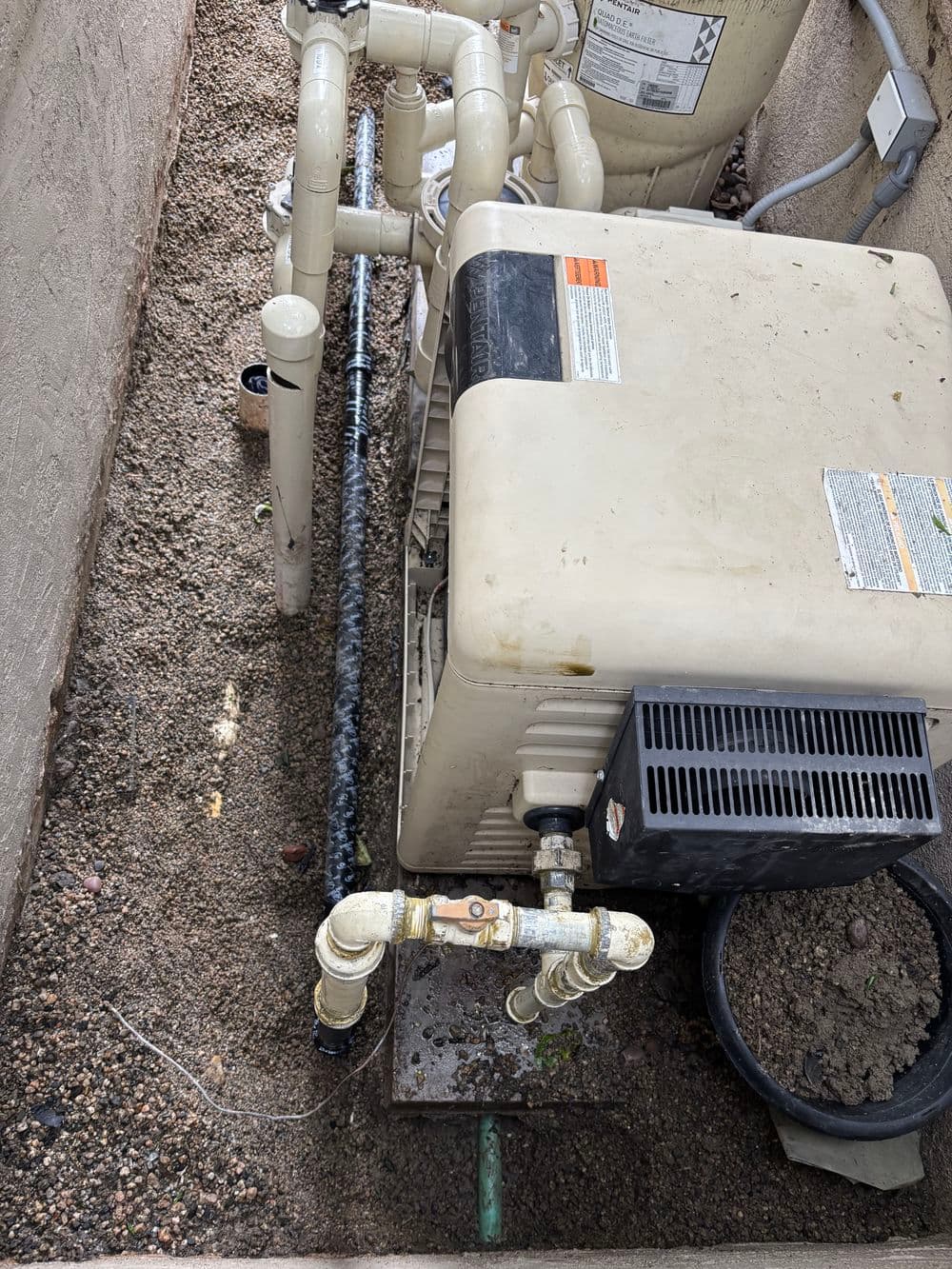 Pool pump system with pipes and equipment in a gravel bed beside a house.