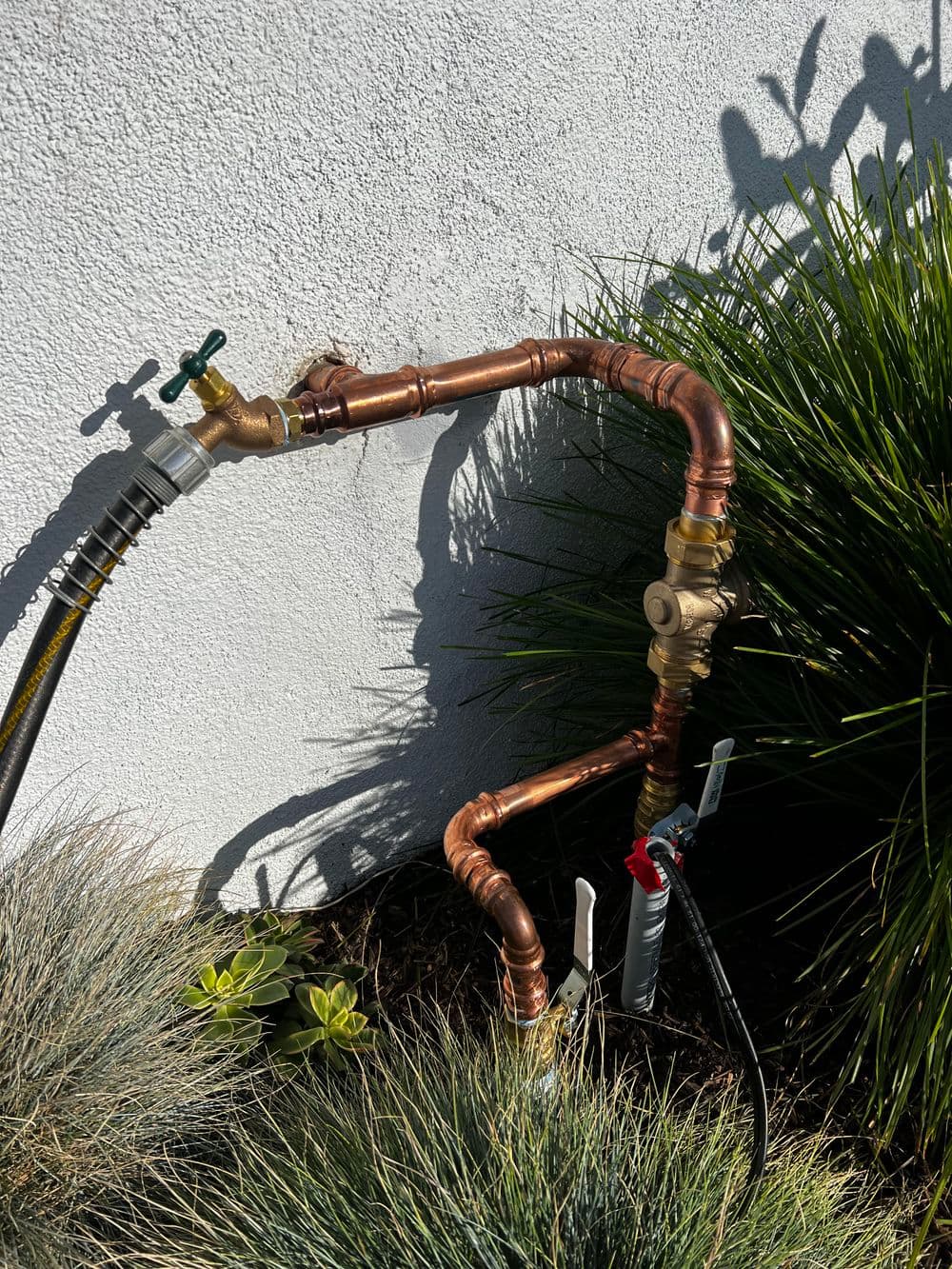Copper plumbing pipes with valves near greenery, showcasing irrigation setup against a wall.