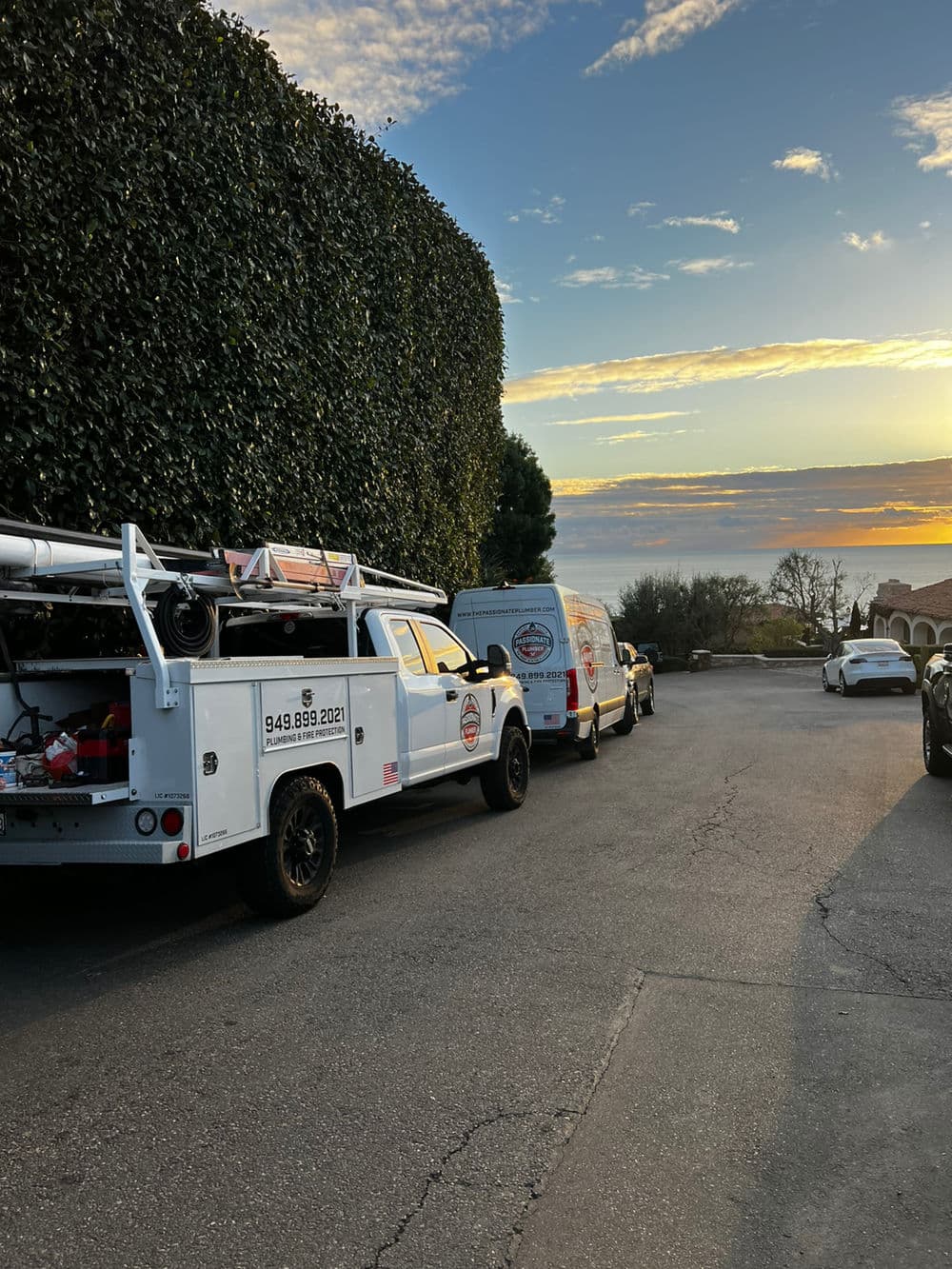 Service trucks parked in a driveway at sunset, surrounded by greenery and a clear sky.